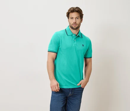 Mann mit einem smaragdgrünen Poloshirt und Jeans, eine Hand in der Hosentasche.