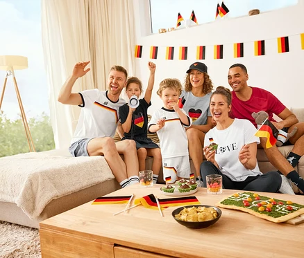 Sechs Personen jubeln auf einem Sofa, tragen Sportshirts, Herz-T-Shirts und Kinder-Trikots. Vor ihnen liegen Cargo-Sweatshorts, eine Sport-Cap und Snacks.