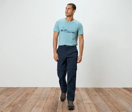 Ein Mann geht auf einem Holzboden, er trägt ein blaues T-Shirt, eine marineblaue Hose und graue Turnschuhe.