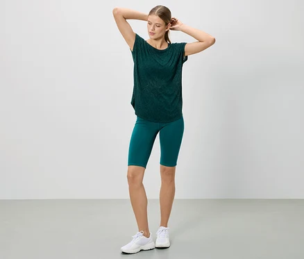 Frau posiert in einem dunkelgrünen T-Shirt und Shorts, dazu weiße Turnschuhe.
