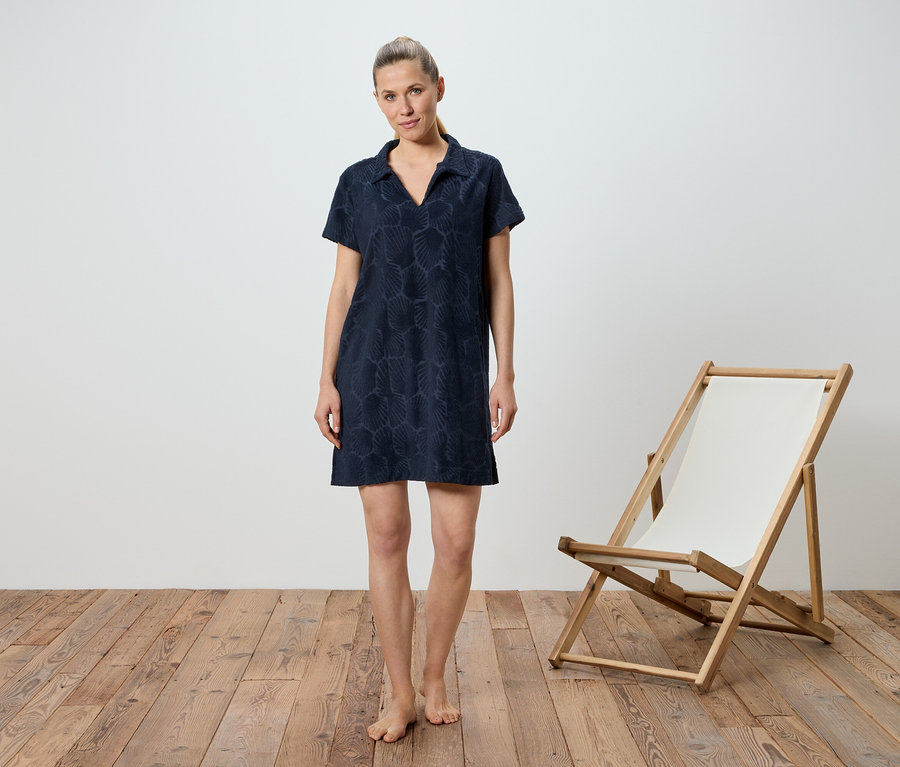 Frau steht barfuß vor einem weißen Hintergrund. Sie trägt ein blaues, kurzärmeliges Kleid. Rechts von ihr steht ein Liegestuhl aus Holz mit weißer Bespannung.
