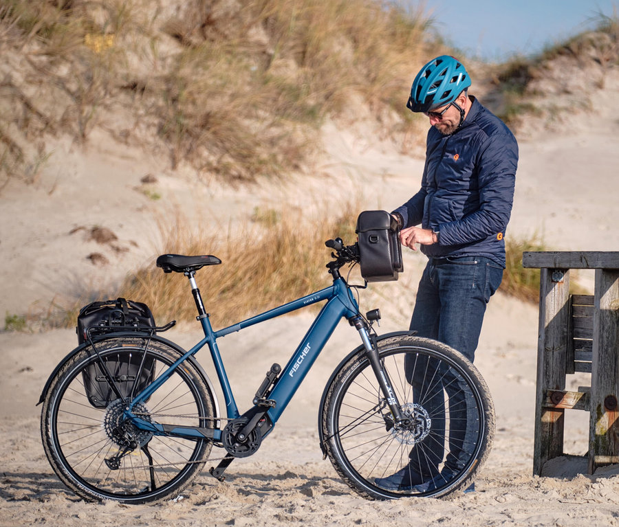 Mann mit blauem FISCHER Fahrradhelm Urban PLUS Chicago neben blauem E-Bike am Strand.