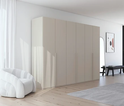 Innenraum mit großem, beigefarbenem Kleiderschrank, weißem Sessel, Spiegel und Bank. Ein grauer Teppich liegt auf dem Holzboden.