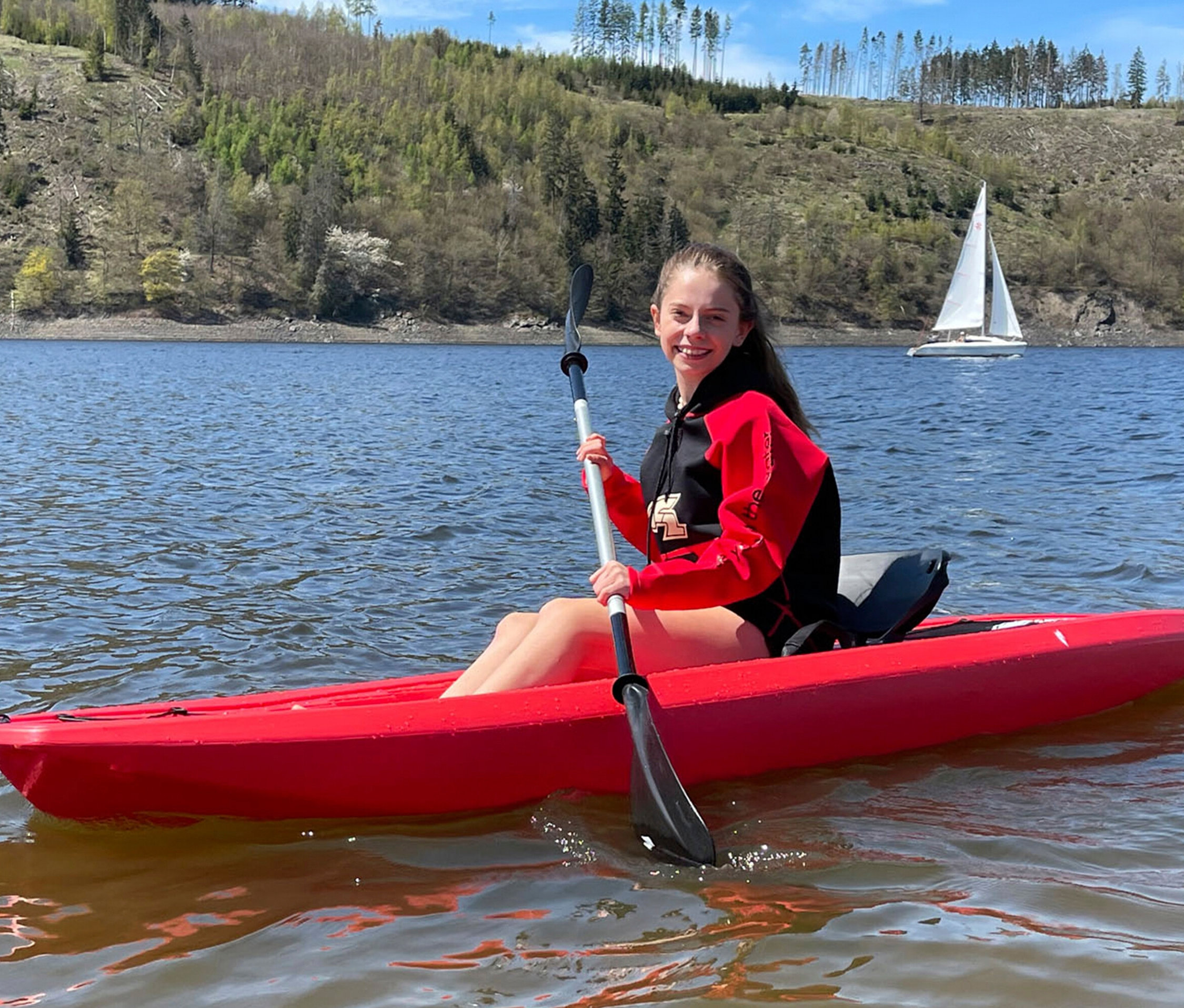 Eine junge Frau paddelt in einem roten Kajak auf einem See. Im Hintergrund sind bewaldete Hügel und ein Segelboot zu sehen.