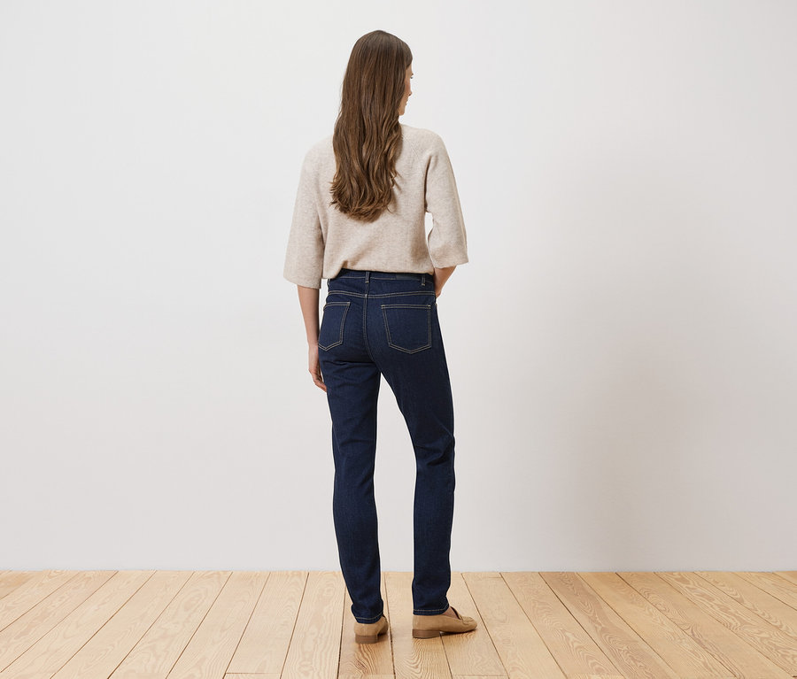 Eine Frau mit langen, braunen Haaren steht mit dem Rücken zur Kamera, trägt eine dunkle Jeans, einen beigen Pullover und beigefarbene Schuhe.