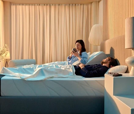 Frau trinkt im Bett, während Mann schläft. Nachttisch mit Lampe und Brille. Vorhang im Hintergrund.