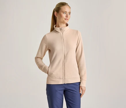 Eine Frau mit blondem Pferdeschwanz trägt eine beigefarbene, gerippte Jacke mit Reißverschluss und blaue Hosen, wobei eine Hand in der Jackentasche steckt.