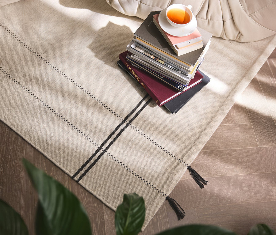 Ein beiger Teppich mit schwarzen Streifen und Fransen liegt auf einem Holzboden, darauf ein Bücherstapel mit einer Tasse Tee.