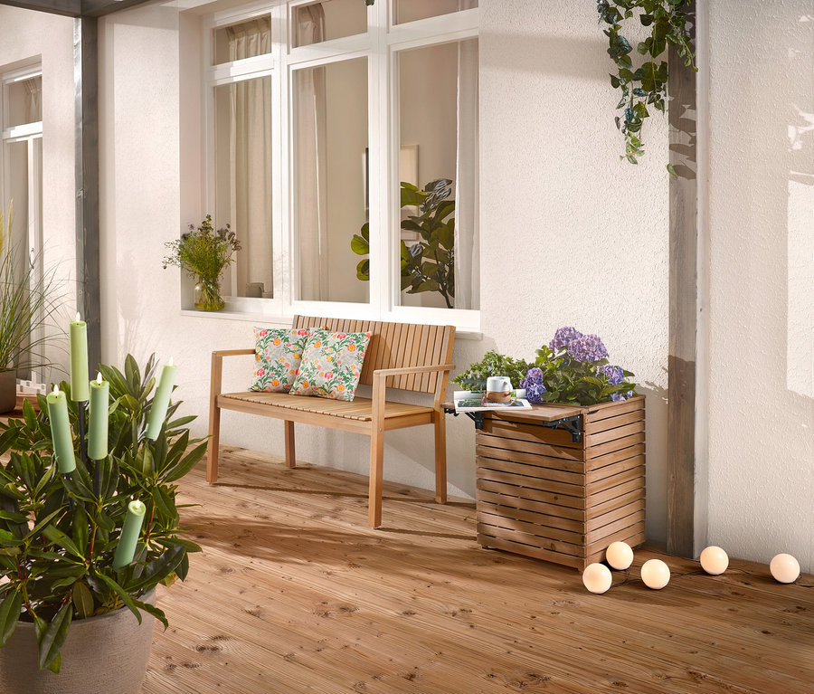 Auf einer Holzterrasse stehen eine Holzbank mit Kissen, ein Pflanzkasten mit Blumen und eine Topfpflanze mit Kerzen.