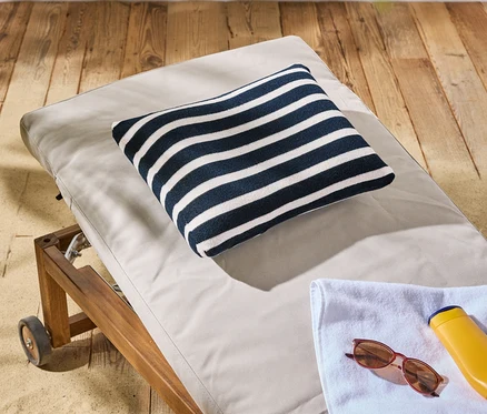 Ein gestreiftes Kissen liegt auf einer Sonnenliege mit Rädern, daneben eine Sonnencreme, eine Sonnenbrille und ein Handtuch.