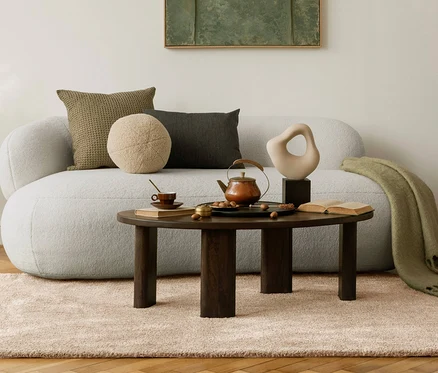 Wohnzimmer mit beigem Teppich »Jane« und Sofa mit Kissen. Auf einem runden Tisch stehen eine Teekanne, eine Tasse und Deko.