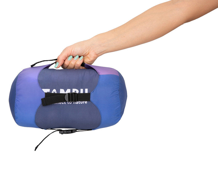 Eine Hand hält einen lila-blauen Schlafsack mit schwarzem Gurt und Kordelzug vor einem weißen Hintergrund.