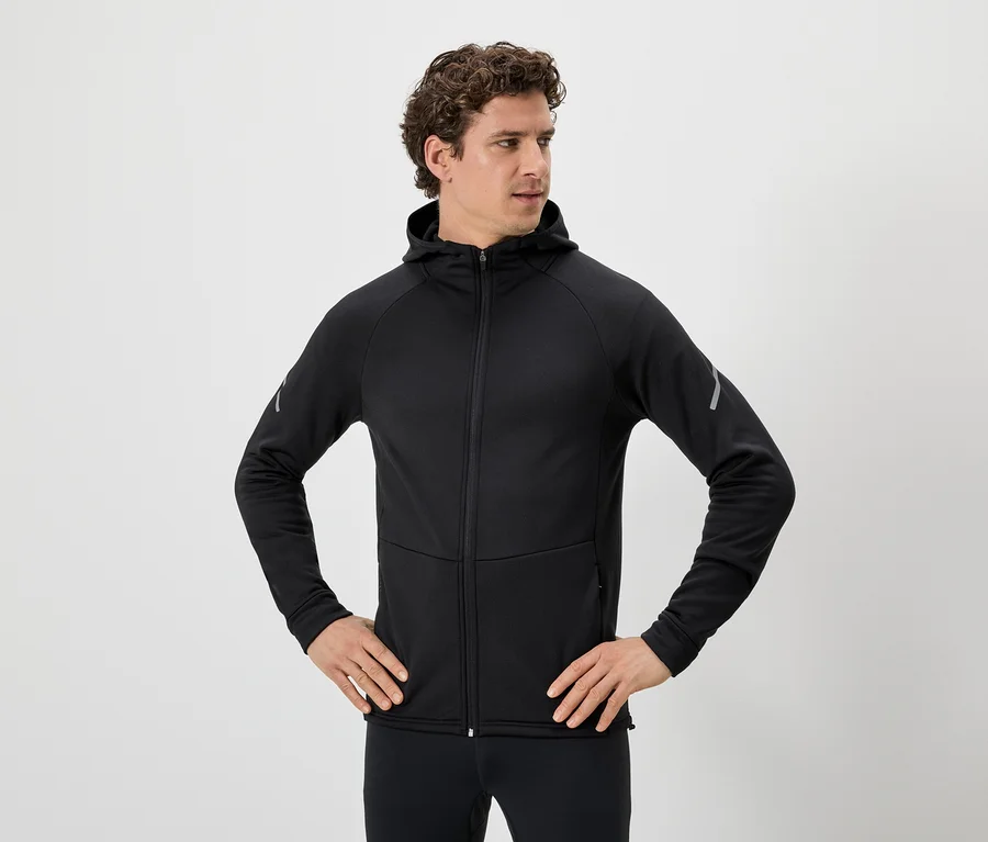 Mann mit lockigem Haar, schwarzer Kapuzenjacke mit Reißverschluss und schwarzen Leggings, die Hände in den Hüften.
