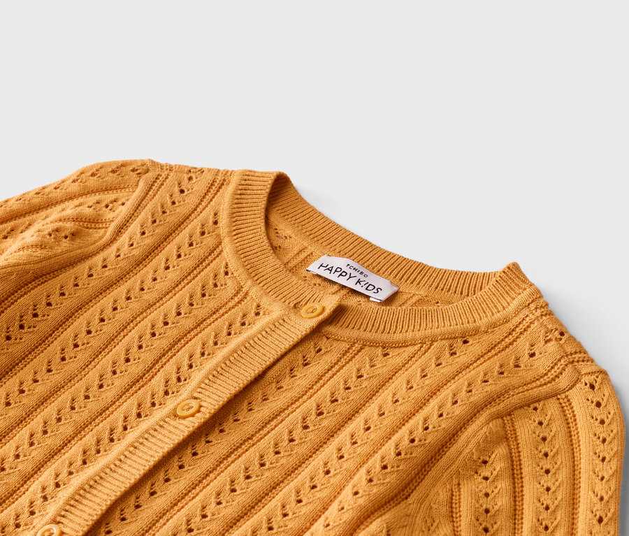 Nahaufnahme einer gelben Strickjacke mit Knöpfen und Lochmuster. Ein Etikett mit der Aufschrift "TCHIBO HAPPY KIDS" ist sichtbar.