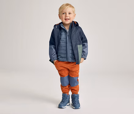 Ein lächelnder Junge mit blonder Kurzhaarfrisur trägt eine blaue Jacke, eine blaue Steppweste, orangefarbene Hosen und blaue Stiefel.