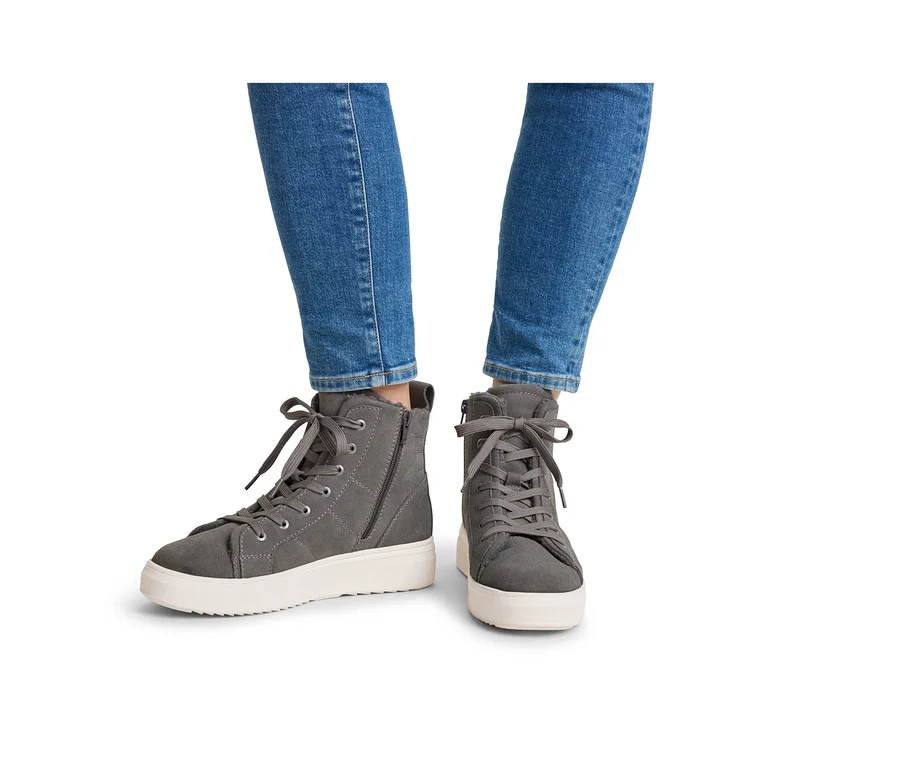 Beine einer Person in blauen Jeans und grauen High-Top-Sneakern mit weißen Sohlen, Schnürsenkeln und seitlichen Reißverschlüssen.