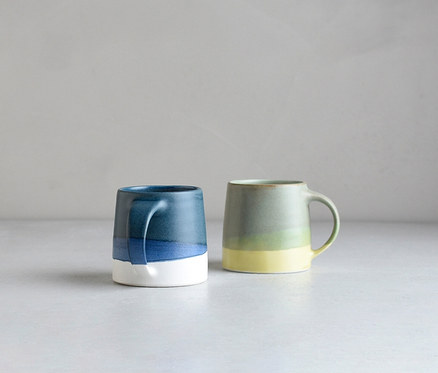 Zwei Keramiktassen stehen auf einer hellgrauen Oberfläche. Die linke Tasse ist oben dunkelblau, geht in Hellblau über und ist unten weiß. Die rechte Tasse ist oben hellgrün, geht in Gelbgrün über und ist unten gelb.