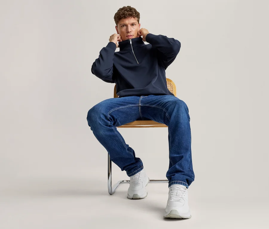 Ein Mann sitzt auf einem Stuhl, richtet den Kragen seines dunkelblauen Half-Zip-Sweatshirts und trägt blaue Jeans sowie weiße Sneaker.