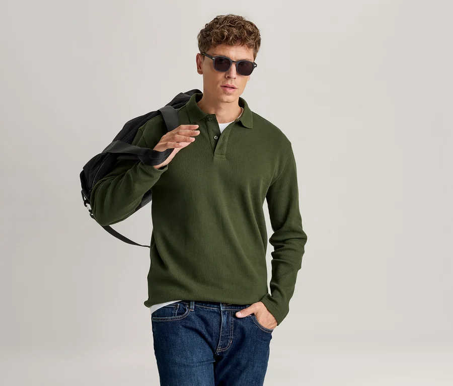 Mann in grünem Langarm-Poloshirt, blauer Jeans und Sonnenbrille, mit schwarzer Tasche über der Schulter.