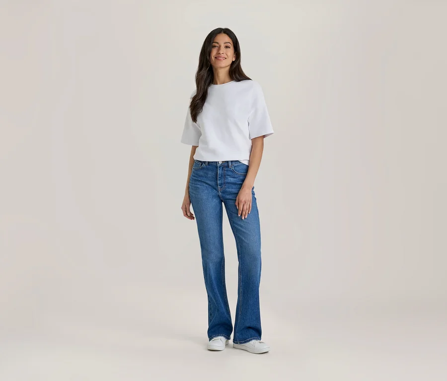 Eine Frau mit langen dunklen Haaren, die ein weißes Kurzarm-Oberteil und eine blaue Schlagjeans trägt, steht nach vorne blickend und lächelt. Sie trägt auch weiße Turnschuhe.