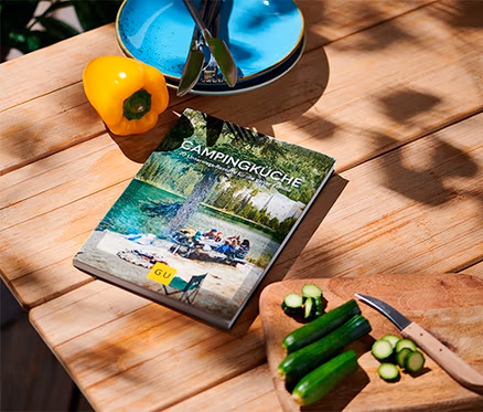 Auf einem Holztisch liegen das Buch »Campingküche«, eine gelbe Paprika, Teller und geschnittene Zucchini.