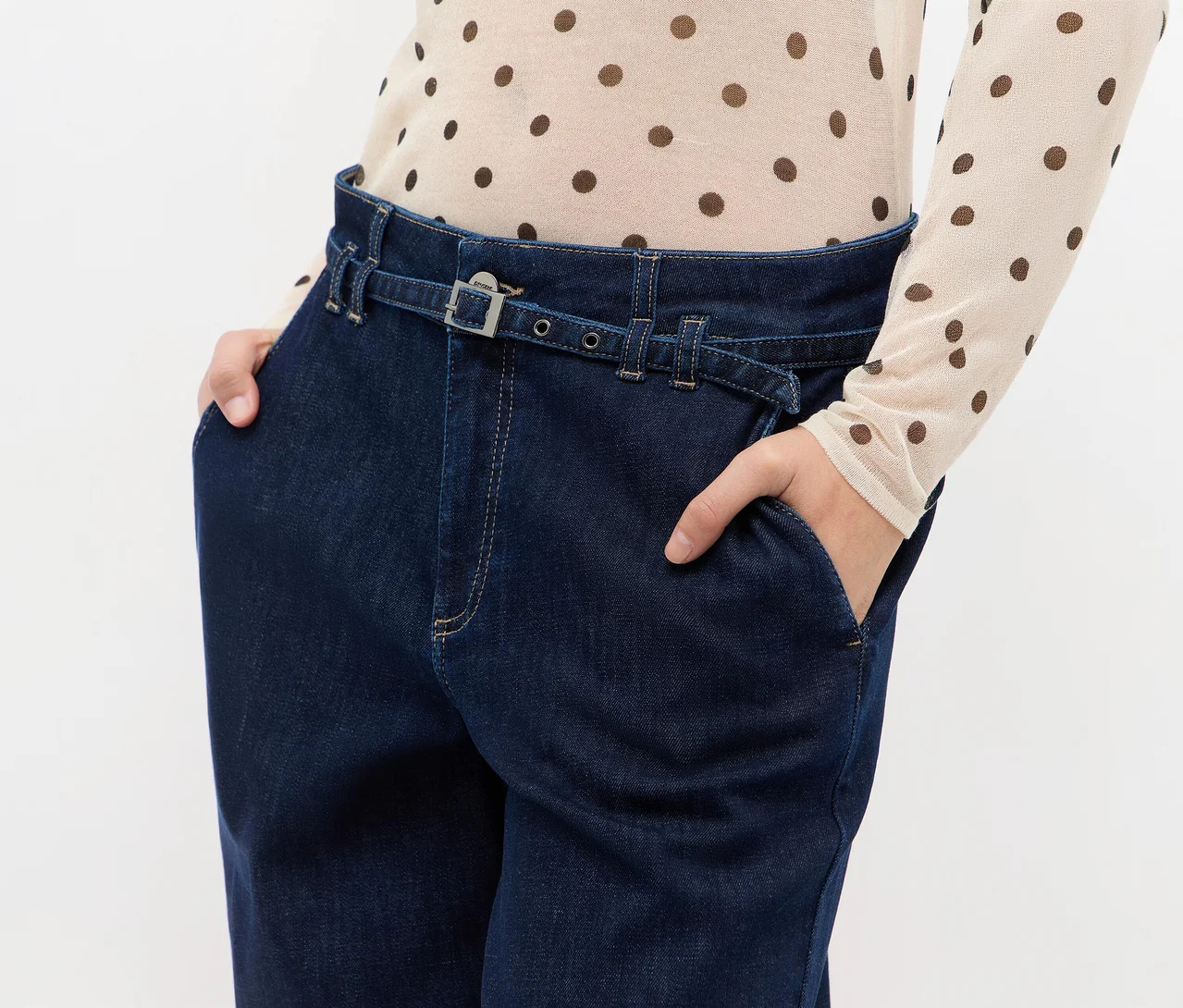Person trägt dunkelblaue Jeans mit integriertem Gürtel und ein helles, gepunktetes Oberteil. Die Hände stecken in den Hosentaschen.