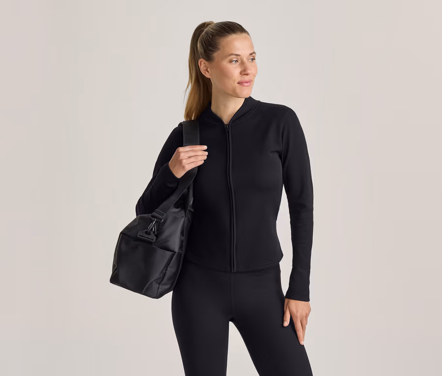 Frau in schwarzer Sportkleidung, bestehend aus einem langärmeligen Oberteil mit Reißverschluss und Leggings, trägt eine schwarze Sporttasche über der linken Schulter.