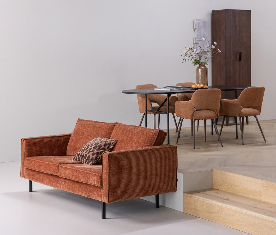 Rostfarbenes Cordsofa mit Leopardenmusterkissen; im Hintergrund ein schwarzer ovaler Esstisch mit vier braunen Polsterstühlen, darauf eine Vase mit Blumen und Gebäck, daneben ein dunkler Holzschrank und eine erhöhte Plattform mit Stufen.