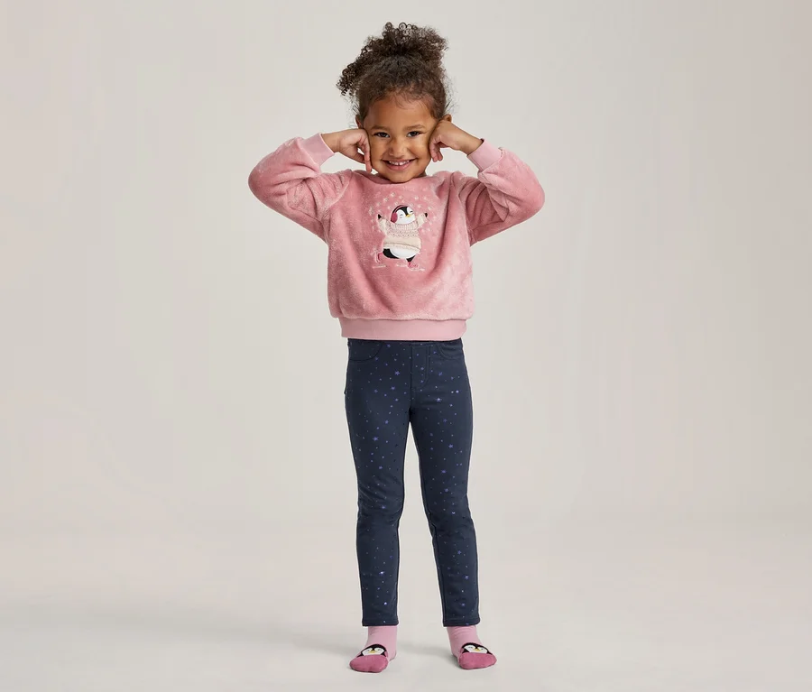 Ein lächelndes kleines Mädchen mit lockigem Haar, das die Hände an die Ohren hält, trägt ein rosa flauschiges Oberteil mit Pinguinmotiv und dunkelblaue Leggings mit Sternenmuster.