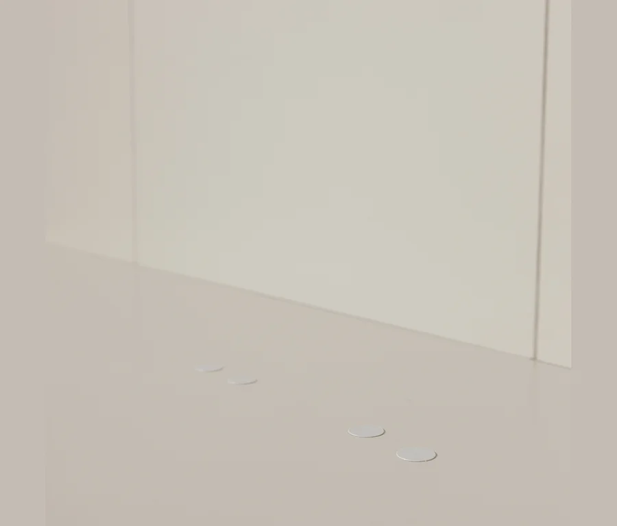 Vier kleine, runde, hellgraue Klebepunkte auf einer hellbeigen Oberfläche, mit einer passenden hellbeigen Wand im Hintergrund.