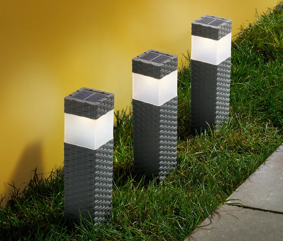 Drei Solarsäulen stehen im Gras neben einem Gehweg.