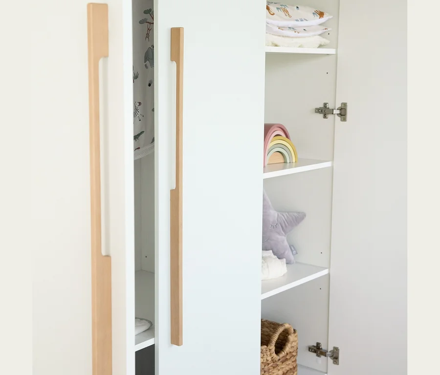 Ein offener weißer Kleiderschrank des Roba 3-teiligen Kinderzimmer-Sets »Lumo« mit Holzgriffen, gefüllt mit einem Holzregenbogen, einem Sternenkissen und Textilien.