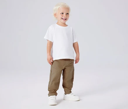 Lächelnder Junge mit weißem T-Shirt und NAME IT Kinder-Twillhose in Khaki und weißen Schuhen.