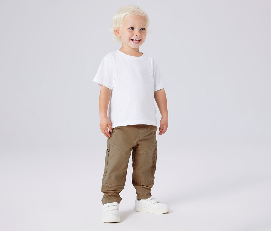 Lächelnder Junge mit weißem T-Shirt und NAME IT Kinder-Twillhose in Khaki und weißen Schuhen.