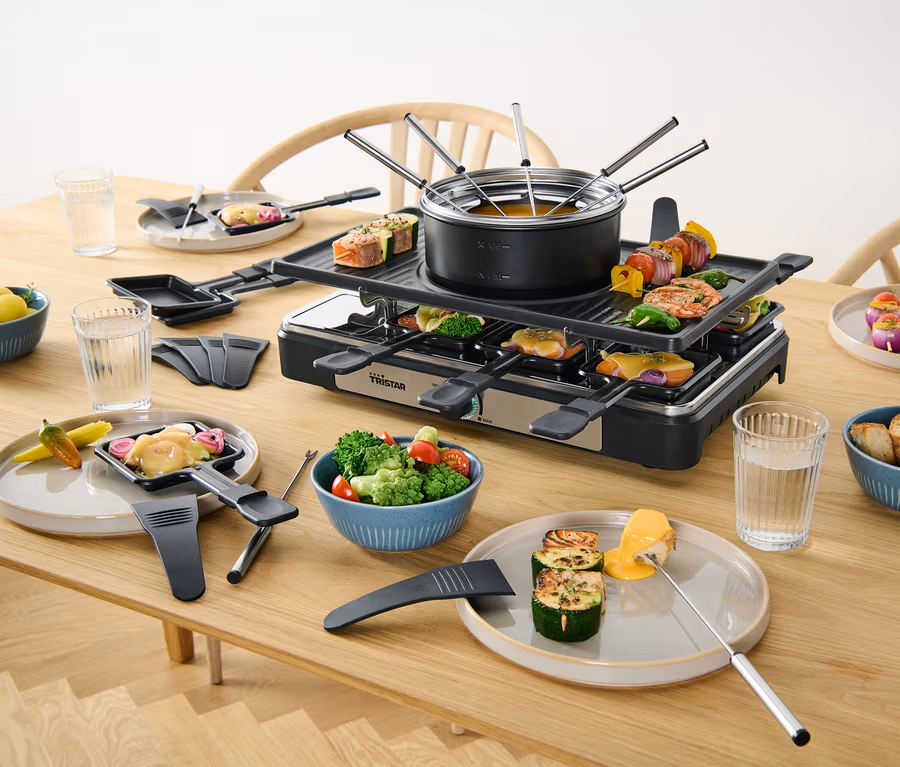 Ein 2-in-1-Raclette-und-Fondue-Gerät steht auf einem Tisch mit Speisen.