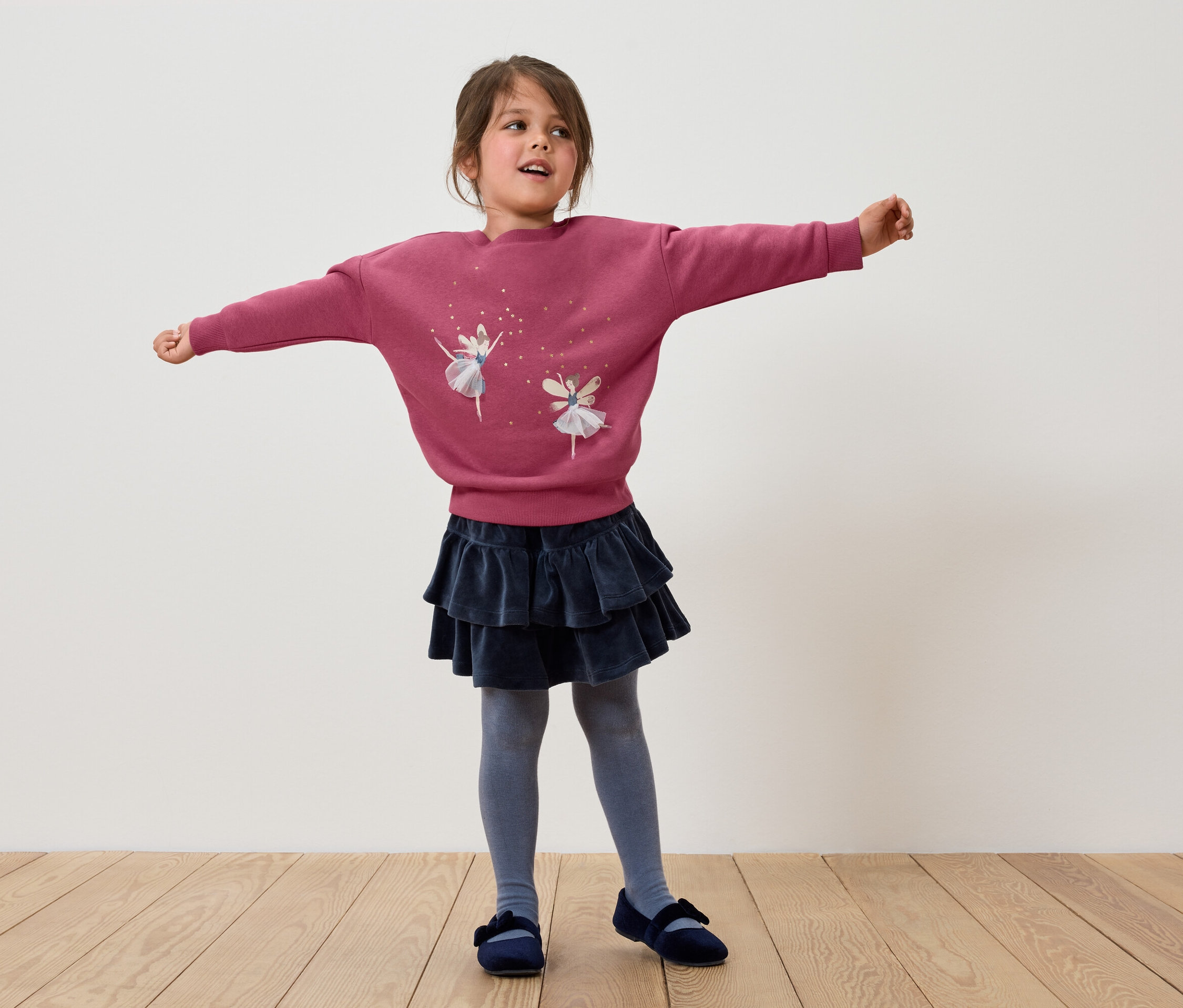 Mädchen steht mit ausgestreckten Armen, trägt ein rosa Sweatshirt mit Ballerinas, einen dunkelblauen Rock mit Rüschen, graue Strumpfhosen und blaue Schuhe.