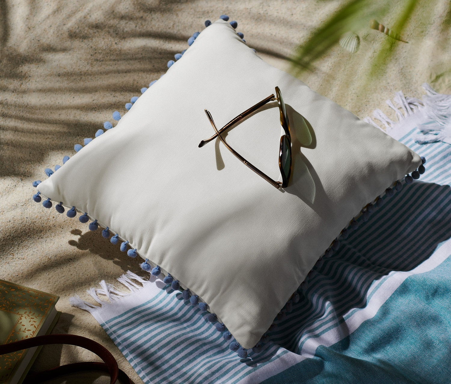 Ein Outdoor-Kissen mit Quasten und eine Sonnenbrille liegen am Strand neben einem gestreiften Strandtuch und einer Strohtasche.