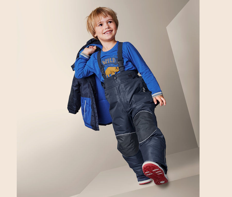 Junge in Schneehose mit recyceltem Material, blauem Shirt und Jacke über der Schulter geht Treppe runter.