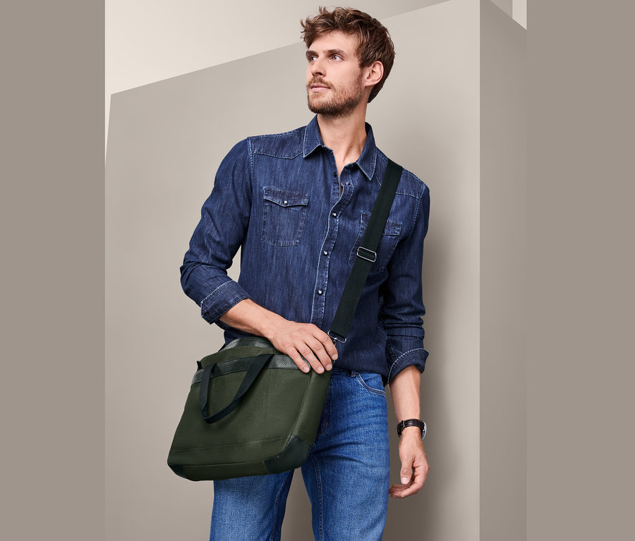 Mann mit braunen Haaren und Bart trägt ein Jeanshemd und Jeans und eine Crossbag über der Schulter.