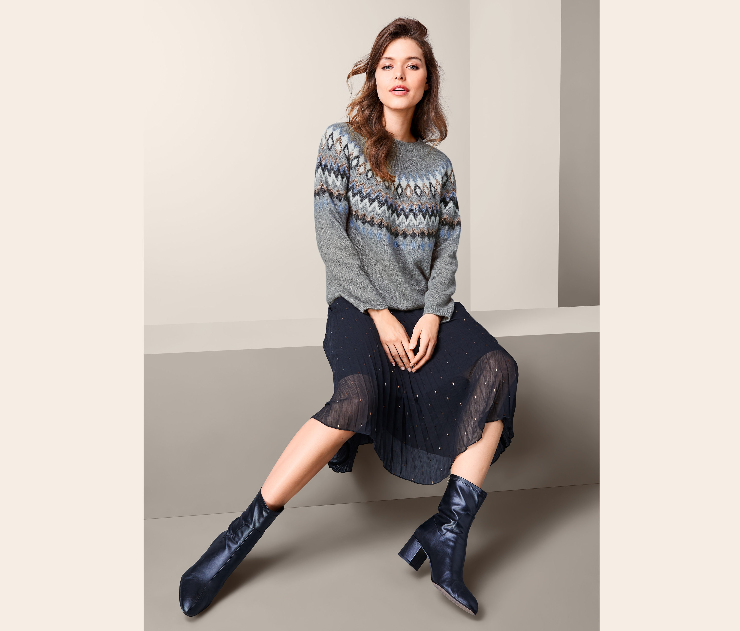 Frau sitzt und trägt einen Norweger-Strickpullover, einen Faltenrock und Lederstiefel.