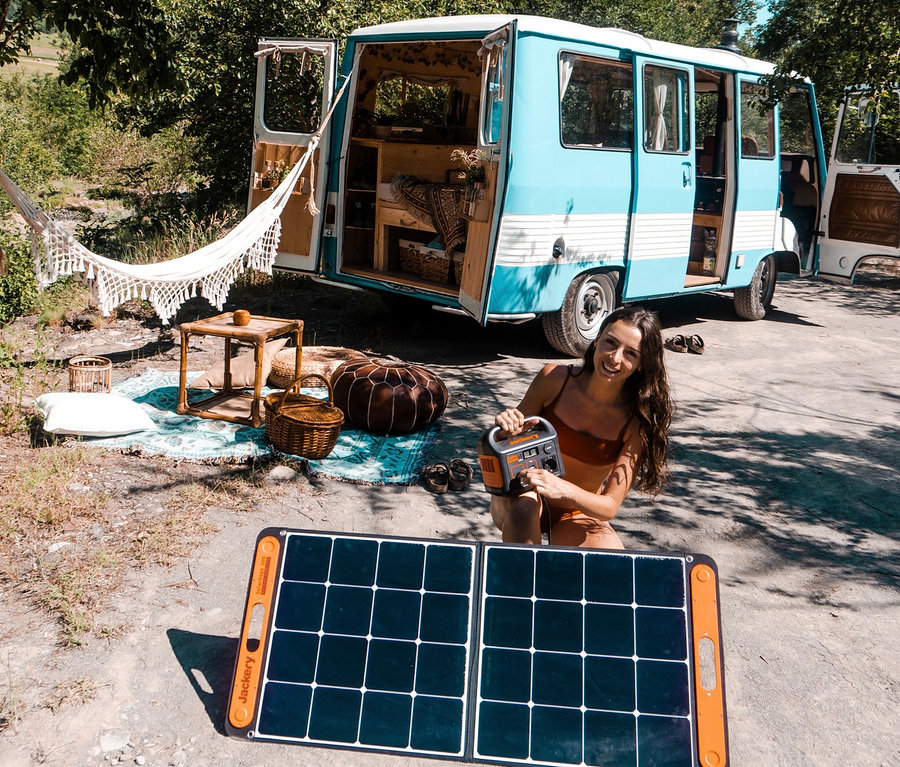 Eine Frau sitzt vor einem blauen Wohnwagen, im Vordergrund ein Solarpanel, sie hält einen Jackery Explorer in der Hand.