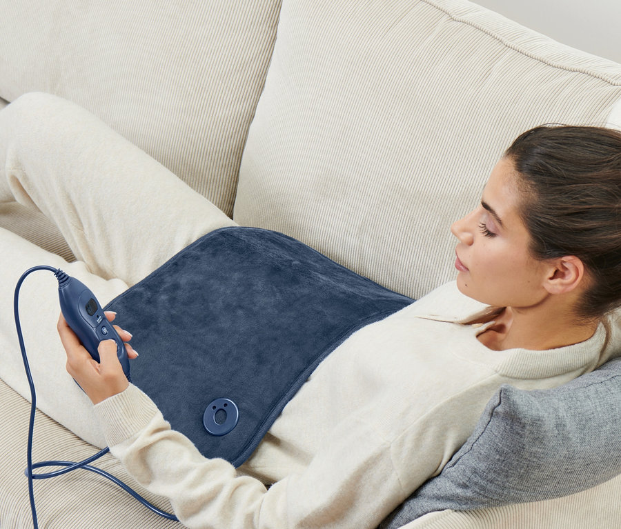 Frau liegt mit kleinem Heizkissen und Fernbedienung auf Sofa.