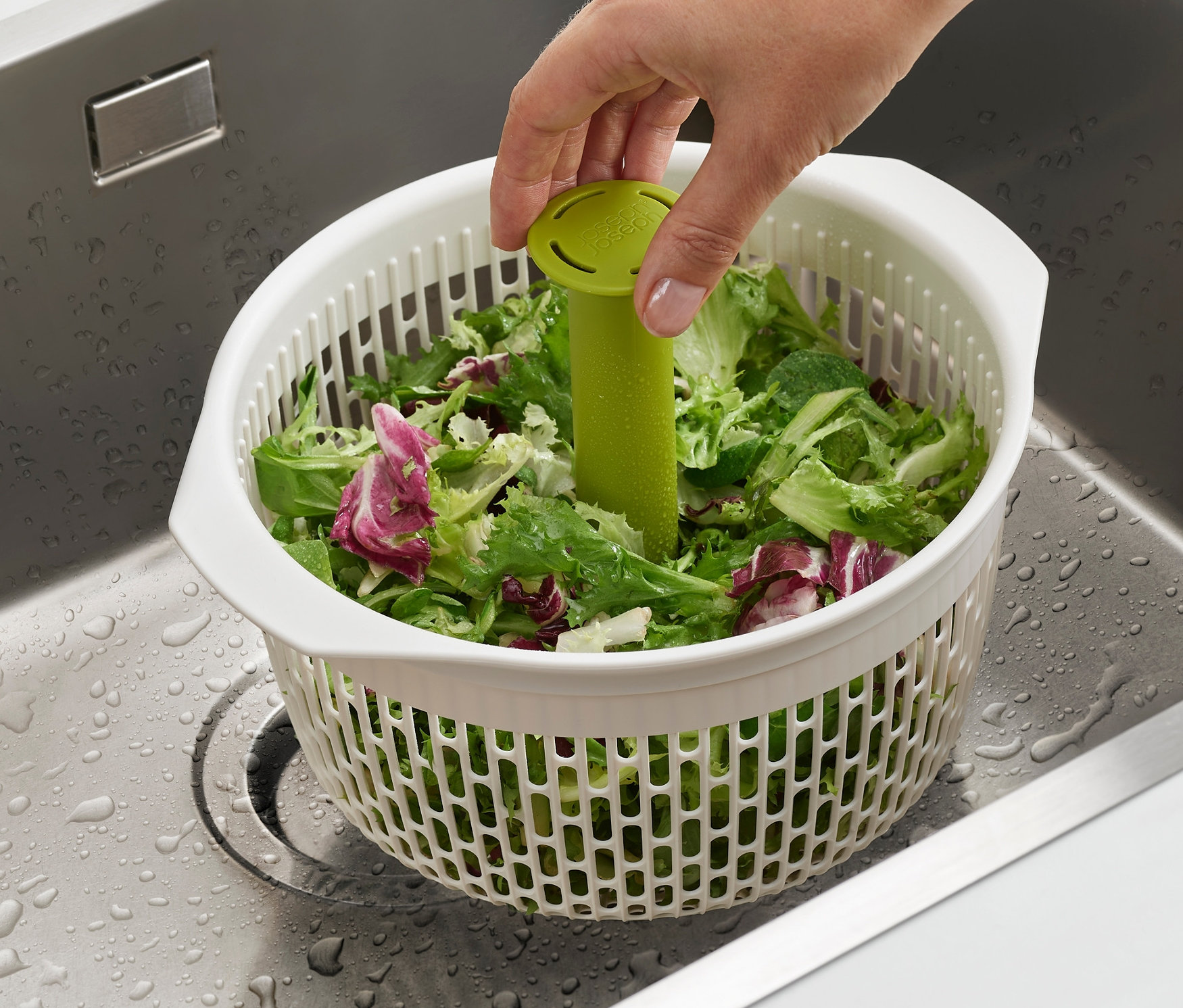 Eine Hand hält einen grünen Presskolben in einer weißen Joseph Joseph 2-in-1-Salatschleuder Spindola™ voller Salat in einem Edelstahlspülbecken.