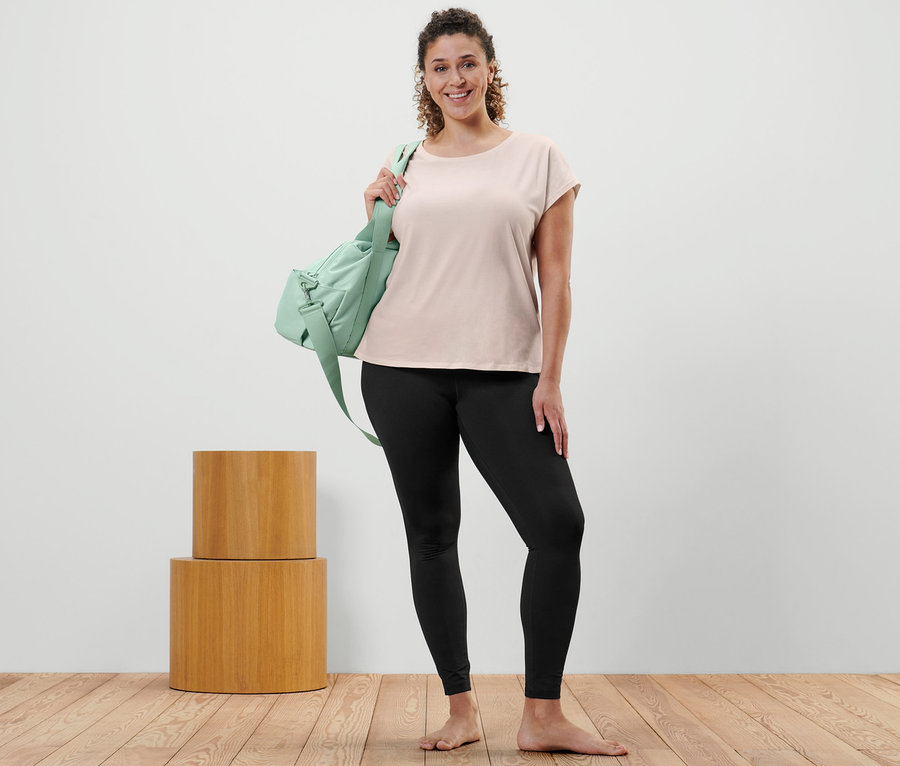 Frau posiert in offwhite Sportshirt, Sporttight und mit grüner Sporttasche über der Schulter.