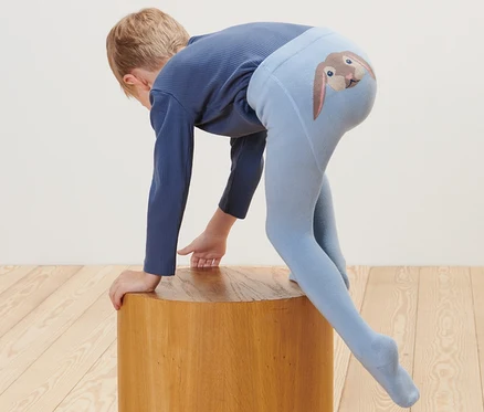 Ein Kind stützt sich auf einen Holzhocker, es trägt eine blaue Strumpfhose mit einem Hasenbild am Po. Auf dem Boden liegt ein Plüschtier. 2 Baby-und-Kleinkind-Strumpfhosen.