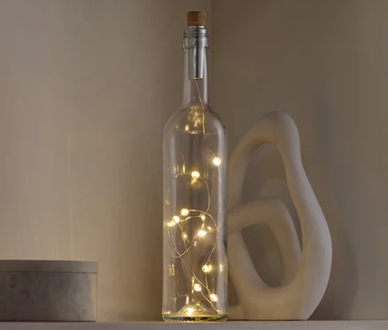 Dekorative Glasflasche mit Korkverschluss und Lichtern steht auf einem Regal neben einer dekorativen Skulptur und einer grauen Schachtel.