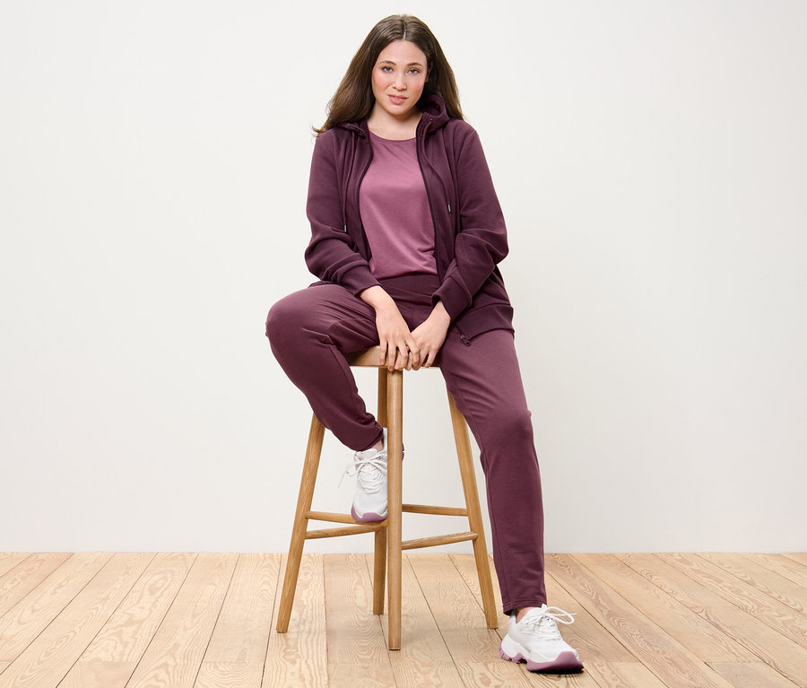 Frau sitzt auf einem Stuhl und trägt eine Sweatjacke, ein langes Shirt in Mauve und eine Sweathose in Pflaume.