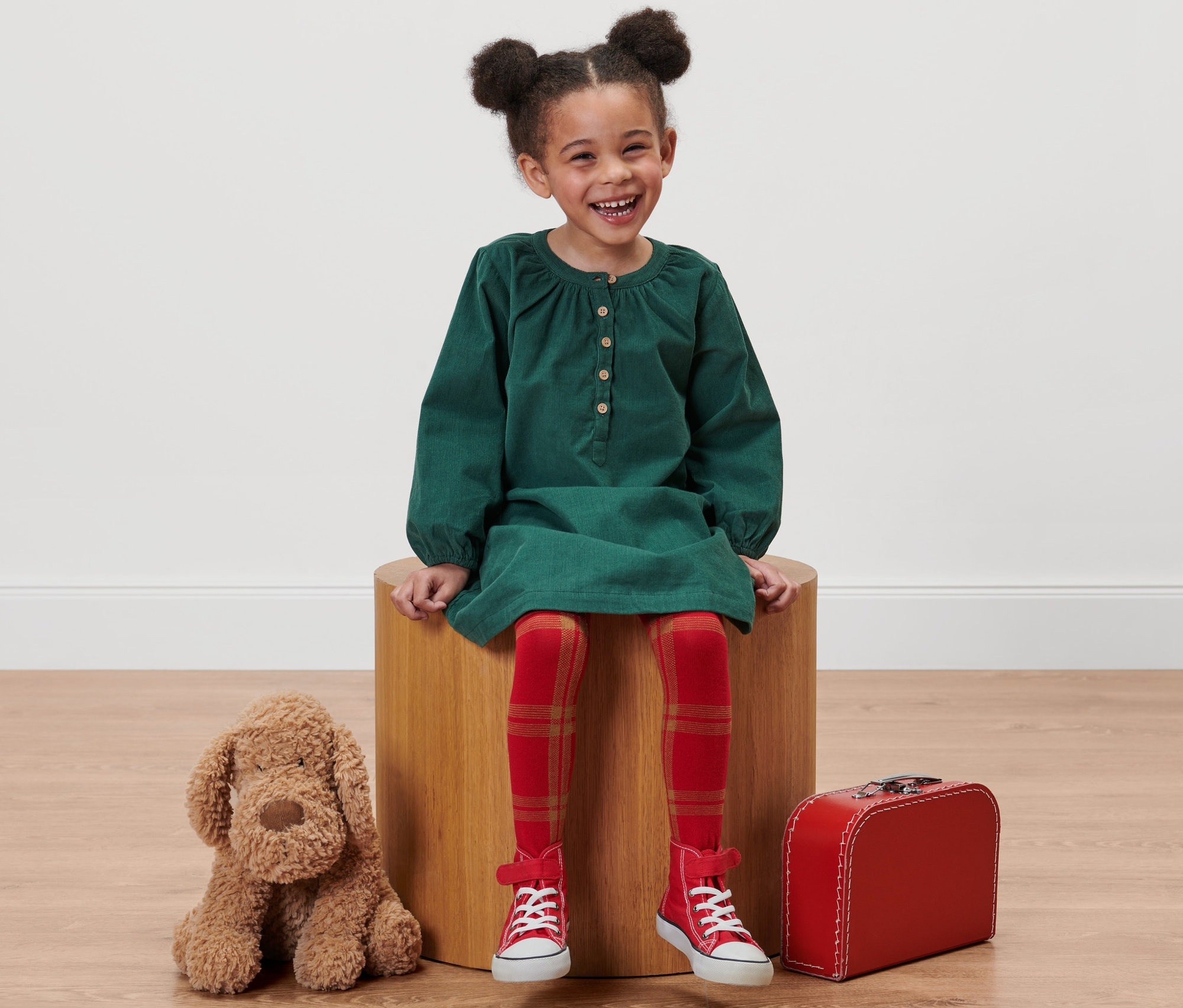 Lächelndes Mädchen sitzt auf einem Holzschemel, trägt ein grünes Kleid, rot-karierte Strumpfhosen und rote Turnschuhe. Neben ihr liegen ein Plüschhund und ein roter Koffer.