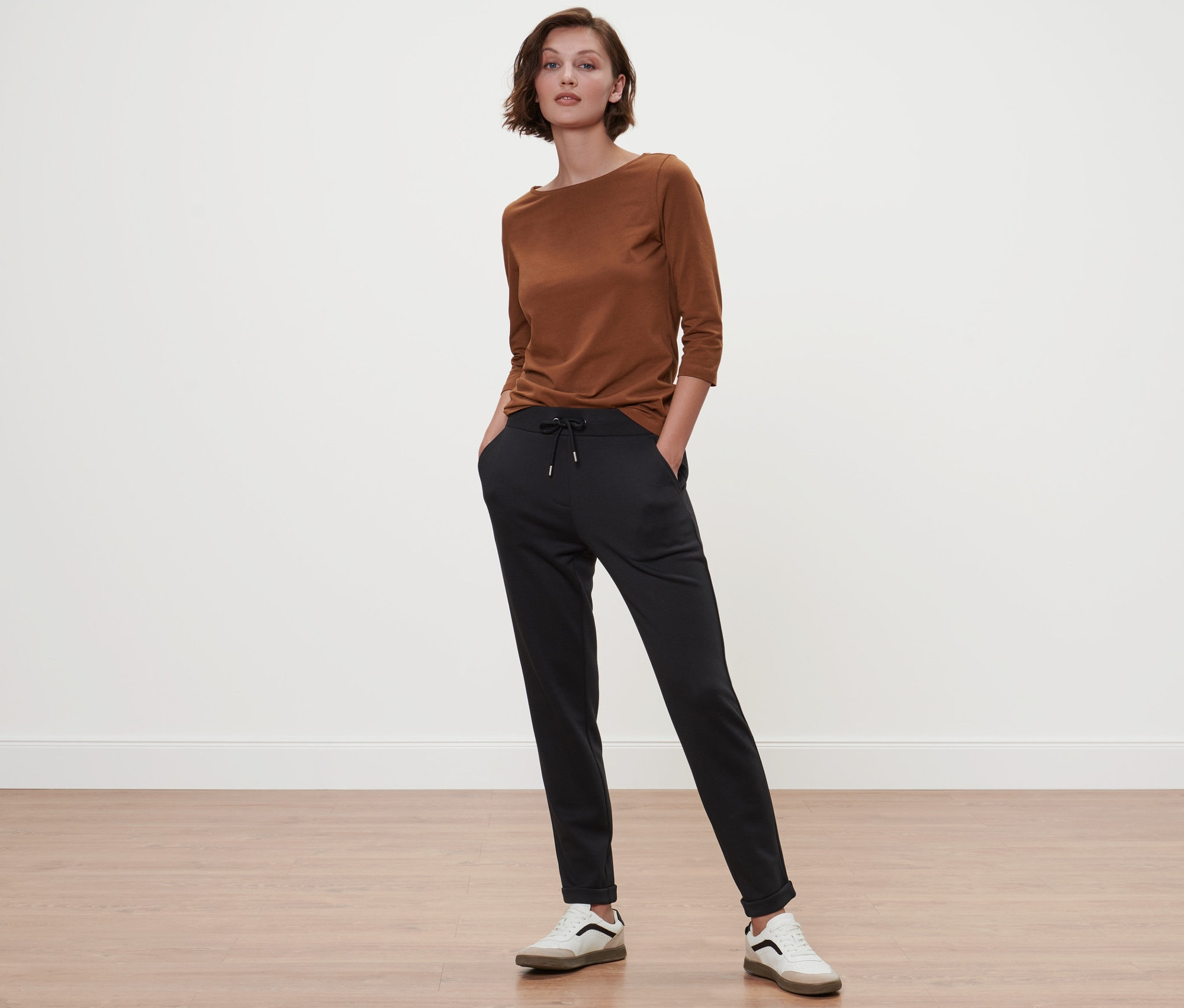 Frau steht mit Händen in den Taschen einer schwarzen Hose und trägt ein cognacfarbenes Shirt mit 3/4-Arm.