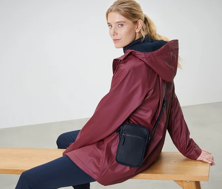 Blonde Frau sitzt auf einer Bank mit einer bordeauxroten Jacke und einer schwarzen Regen-Umhängetasche.